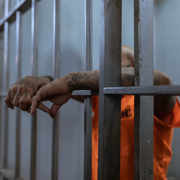 A tattooed inmate's arms rest on jail cell bars, wearing an orange uniform inside a prison.