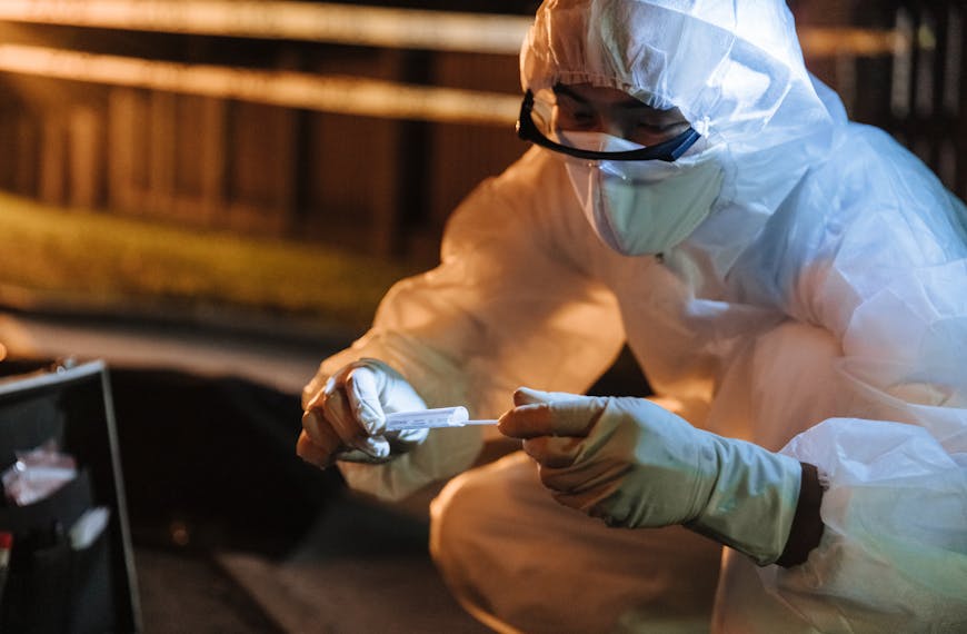 A forensic investigator in protective gear collects evidence outdoors at night.