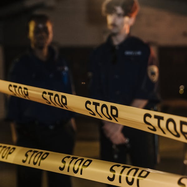 Two police officers standing behind a stop tape at night, scene partially illuminated by car headlights.