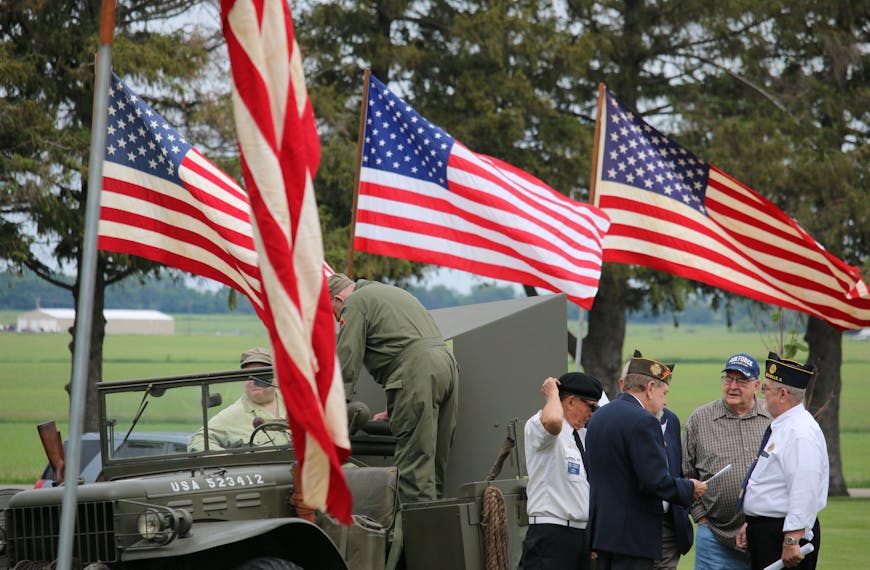 Group of veterans and soldiers by military vehicle with American flags in the background, symbolizing patriotism.
