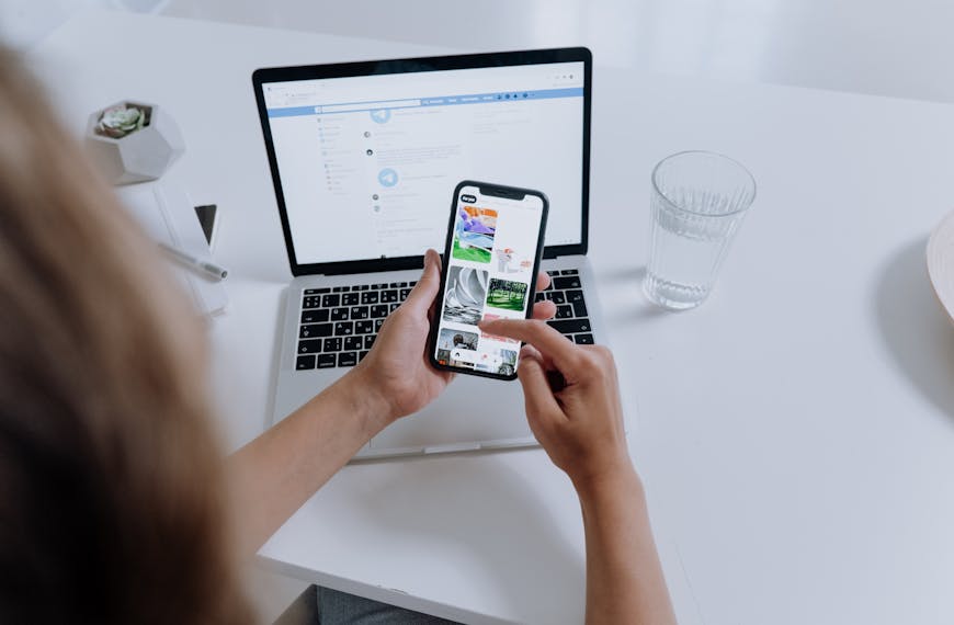 Person using both a smartphone and laptop, multitasking at a modern desk with office plants.